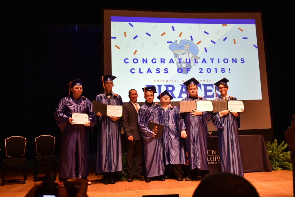 Potential Development graduation ceremony- students wearing blue cap and gown, lined-up holding diplomas on stage